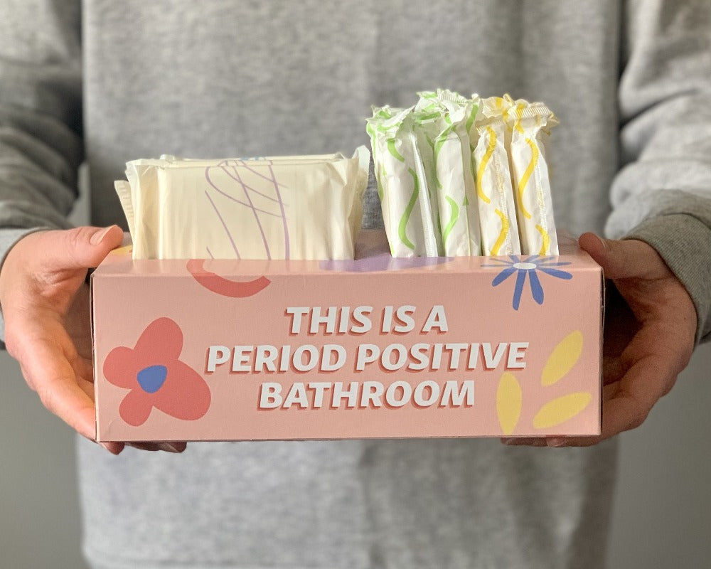 Close up photo of "Your Period Box" on the front it says "This is a period positive bathroom". A person is holding the box up to the camera