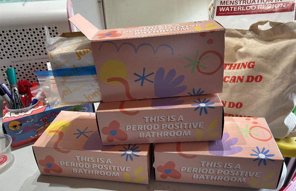 4 period boxes stacked, the sides says This is a period positive Bathroom. Some orgnic pads are sticking out one box. A report called Menstruation in Waterloo Region is in the background. 